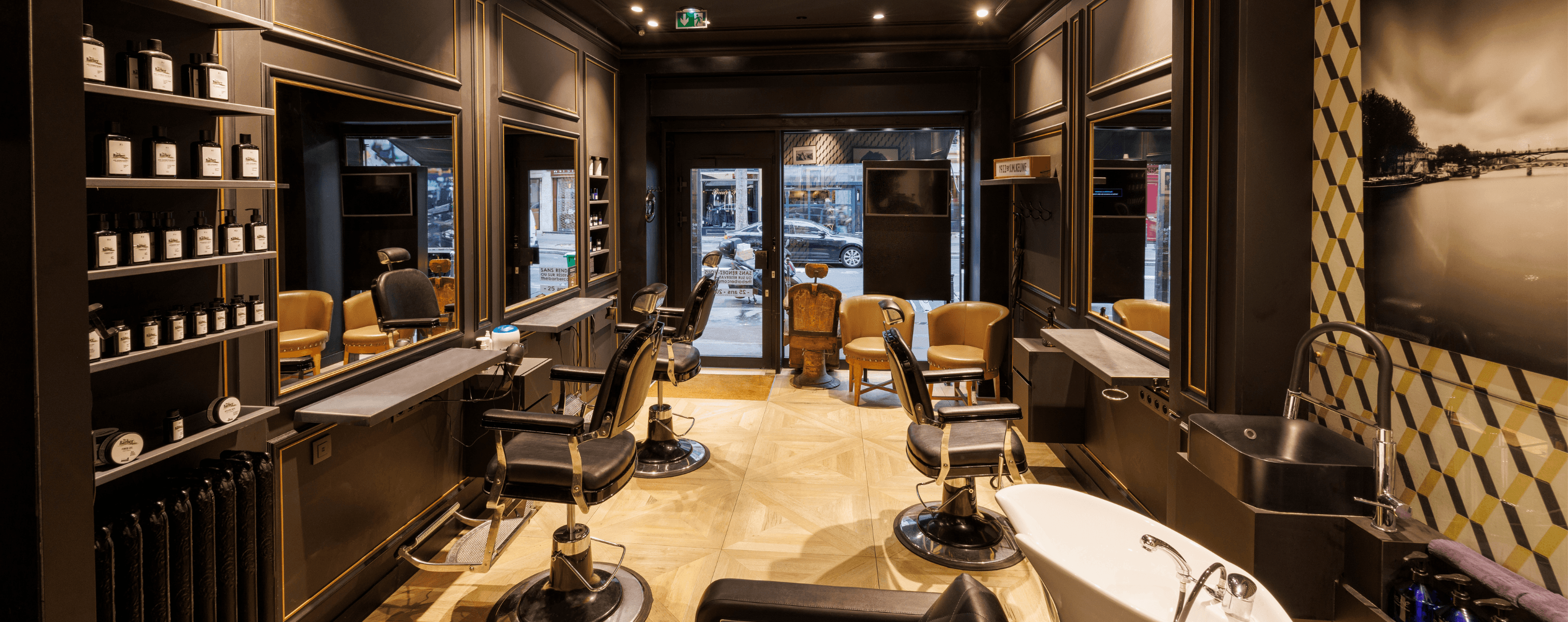 Intérieur élégant d’un salon de coiffure/barbier : plusieurs fauteuils en cuir noir devant de grands miroirs, sol en parquet clair et étagères de produits, avec vue sur la vitrine donnant sur la rue.
